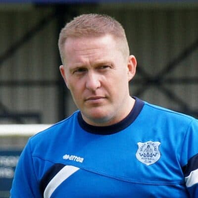 A football player in Bishop Auckland FC kit looking serious, on the pitch during a match at their home stadium.