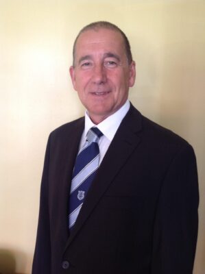 Public domain image of a man in a suit and tie, smiling, with a neutral background, representing a professional or football club official, suitable for SEO on Bishop Auckland FC website.