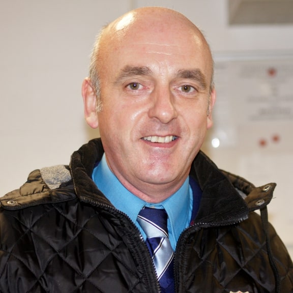 Engaged, smiling man in team staff jacket at Bishop Auckland FC stadium showcasing football community spirit.