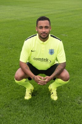 Athletic football player in neon kit crouching on a lush green pitch, representing Bishop Auckland FC, a renowned team in English football.