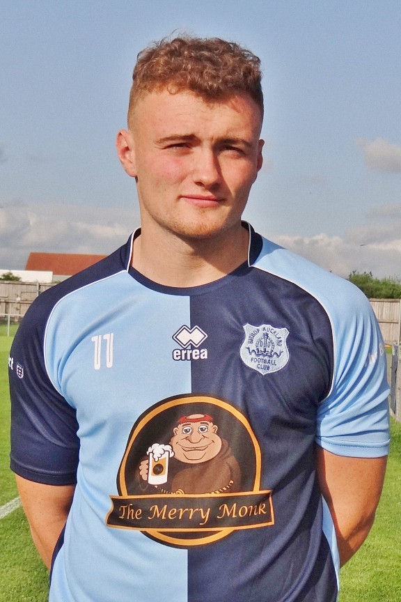 Bishop Auckland FC player in home kit, standing on football pitch during daytime, featuring club badge and sponsorship logo.