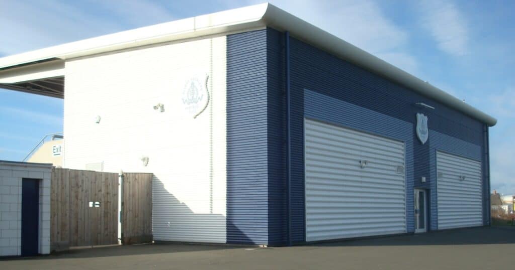Modern football stadium facility at Bishop Auckland FC with blue and white exterior walls, roller shutter doors, and team crest display, supporting local community football activities.