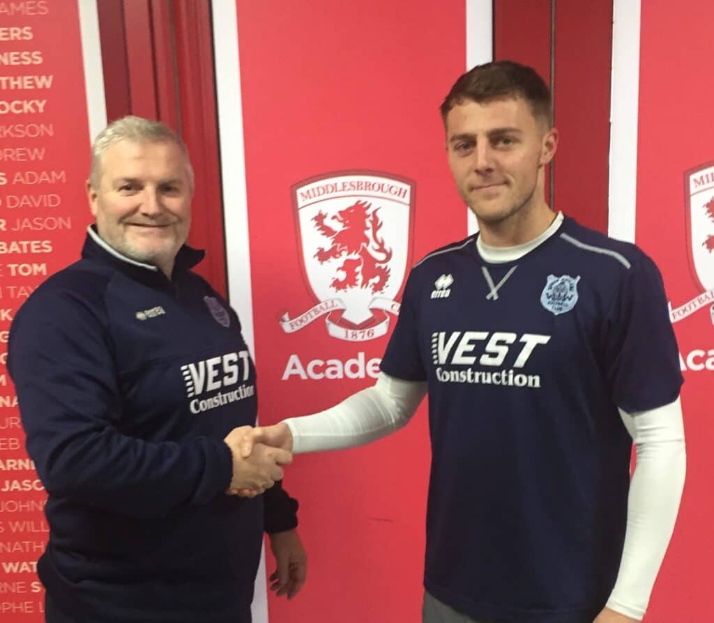 - A man and a young male football player shaking hands at Middlesbrough FC, with team logos in the background, representing Bishop Auckland FC.