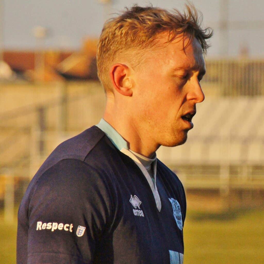 A football player from Bishop Auckland FC with short blonde hair, wearing the team's navy blue kit, standing on the pitch during a match.