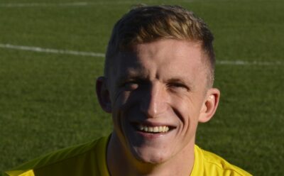 Bishop Auckland FC, football player on the field, smiling in a yellow jersey, representing the historic Northern League football club, supporting local football and community engagement.