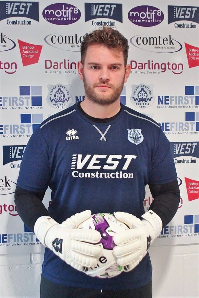 - Goalkeeper in blue football kit holding a purple and white ball in front of a backdrop with sponsorship logos and club branding.