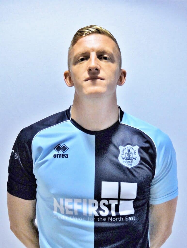 Bright young footballer in Bishop Auckland FC jersey standing against plain background, showcasing the team’s colours and emblem, representing local football club and sporting excellence.