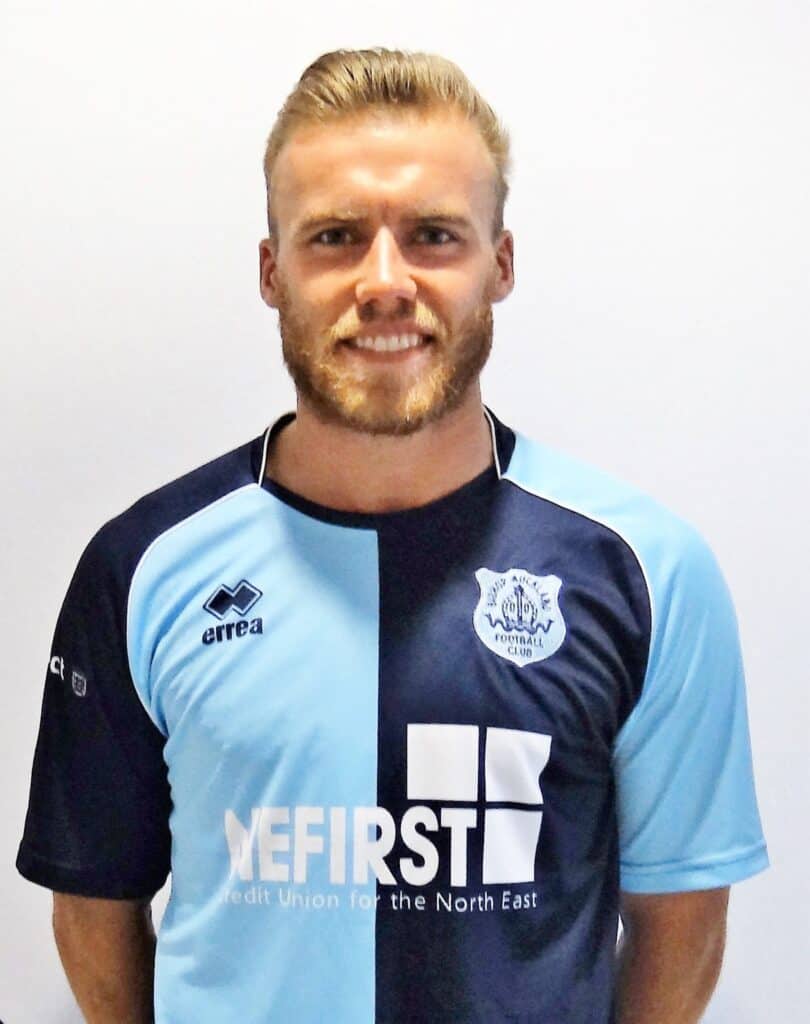 Bright smiling football player in blue and black kit of Bishop Auckland FC, standing against a plain white background. The player is part of the Bishop Auckland football team, showcasing team colours and crest.