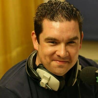 A radio presenter wearing headphones in a BBC studio, smiling at the camera, promoting Bishop Auckland FC with a focus on local football and community support.
