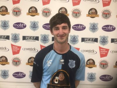 Youth football player in Bishop Auckland FC kit standing in front of a sponsorship backdrop.