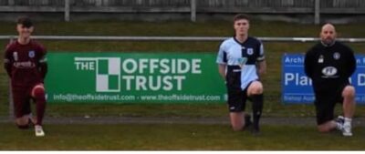 Kneeling football players and coach at Bishop Auckland FC ground, promoting local football club in County Durham, England, with sponsorship banners in the background.