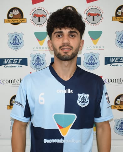 Young male football player wearing Bishop Auckland FC kit, smiling at the camera, standing against a sponsor backdrop, representing the club's team at a promotional event or match.