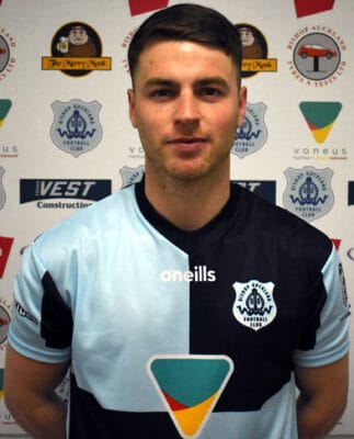 Young footballer in Bishop Auckland FC kit, standing in front of club branding and sponsorship logos, showcasing the team’s player roster and club pride.