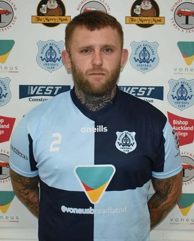 Professional football player in Bishop Auckland FC kit, standing in front of club sponsors backdrop, showcasing local football team and community engagement.