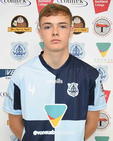 Young Bishop Auckland FC football player standing in front of team sponsorship banners, showcasing club kit and logo, promoting local football clubs in County Durham.