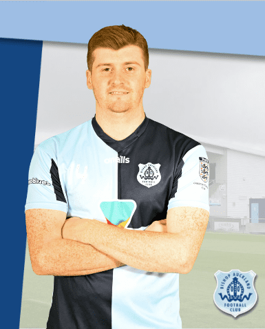 Young male Bishop Auckland FC player in team kit, standing confidently with arms crossed against stadium background.