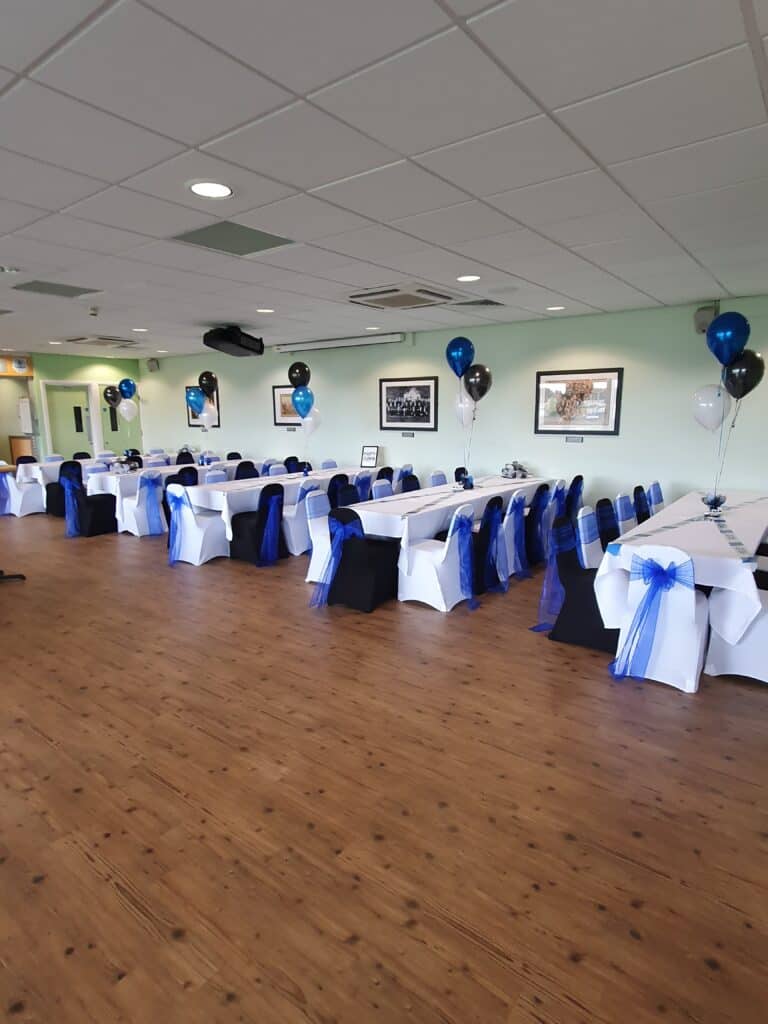 Bishop Auckland FC event hall decorated with white tablecloths, black and blue chair covers, and blue, black, and white balloons for a celebration or gathering.