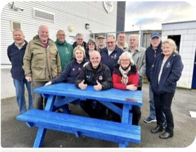 A group of diverse supporters and staff at Bishop Auckland FC posing outdoors with a bright blue picnic table.