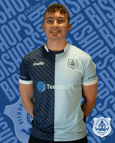 Striker wearing Bishop Auckland FC home kit, standing confidently in front of branded background, representing the football club’s commitment to local sports excellence.