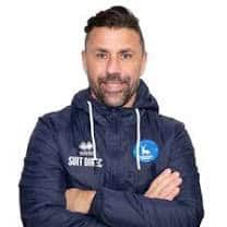 Bishop Auckland FC team staff member wearing club branded jacket, standing with arms crossed, smiling confidently at a sports event.