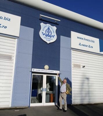 Bishop Auckland FC stadium exterior entrance with club signage and a man pointing at the club badge in bright sunlight.