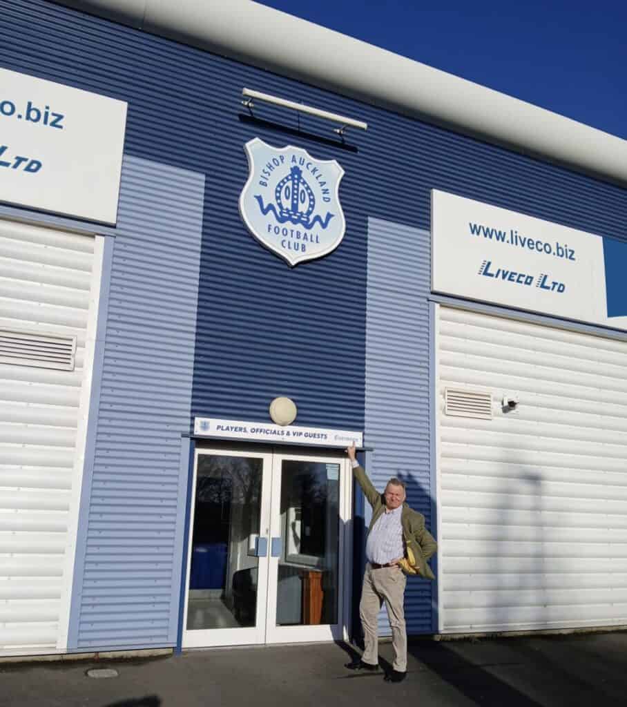 Bishop Auckland FC stadium exterior entrance with club signage and a man pointing at the club badge in bright sunlight.
