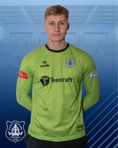 Young football player in green goalkeeper jersey representing Bishop Auckland FC, standing indoors with arms behind back, showcasing team logo and sponsor.