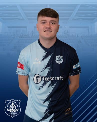 Portrait of a Bishop Auckland FC football player in team kit, standing inside the club's stadium.