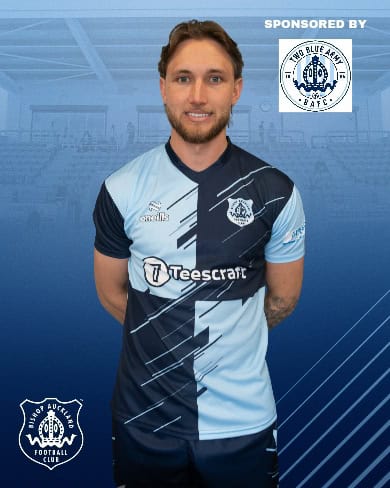 Bishop Auckland FC football jersey featuring blue and navy design with sponsor logos, displayed on a mannequin in a stadium setting.