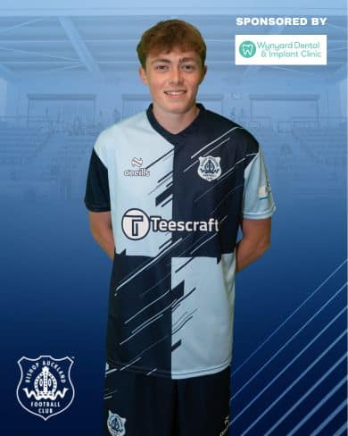 Young Bishop Auckland FC player in team kit, smiling, standing in stadium background, promoting local football club and community engagement.